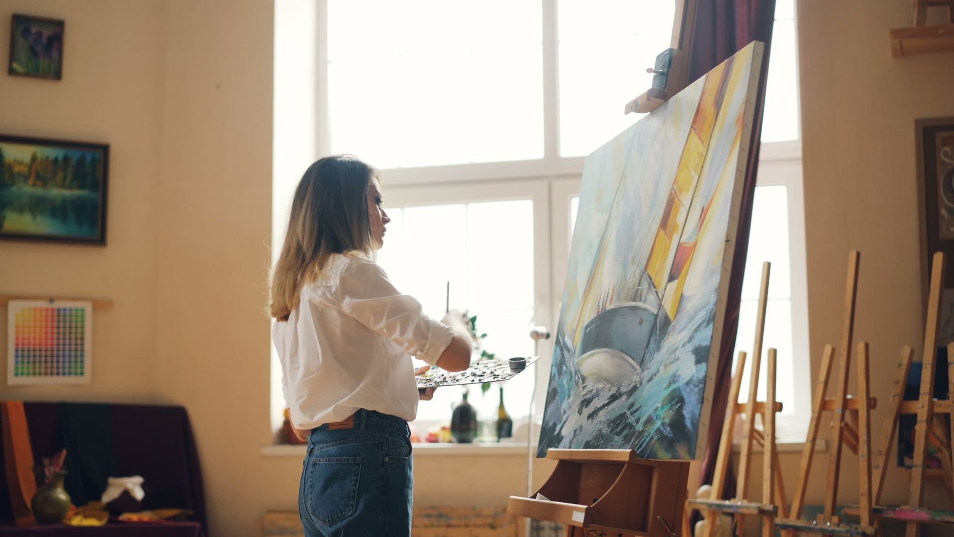 Artist painting a vibrant sailboat on canvas in a sunlit studio.
